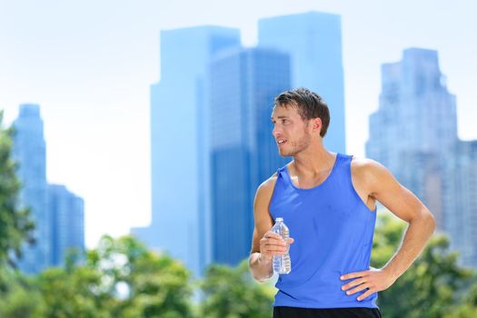 Sport man drinking water bottle in New York City. Male runner sweaty and thirsty after run in Central Park, NYC, Manhattan, with urban buildings skyline in the background.