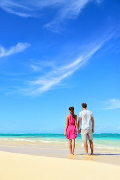 Beach vacation couple relaxing on summer holidays. Young people standing from behind holding hands looking at the ocean, vertical crop with a lot of copy-space in the blue sky. Travel concept.