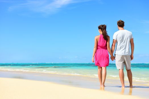 Summer holidays - couple on tropical beach vacation standing in white sand relaxing looking at ocean view. Romantic young adults holding hands in beachwear with pink dress and surf shorts in love.