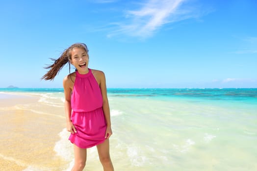 Asian Chinese tourist woman having fun on beach. Young mixed race female adult playing in water wearing pink halter sundress laughing during summer travel holidays. Tropical sunny destination.