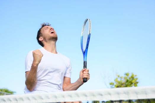 Tennis player man winning cheering celebrating victory. Winner man happy in celebration of success and win. Fit male athlete on tennis court outdoors holding tennis racket in triumph by the net.