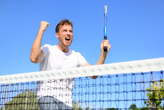 Tennis player celebrating victory. Winning cheering man happy in celebration of success and win. Fit male athlete winner on tennis court outdoors holding tennis racket in triumph by the net.