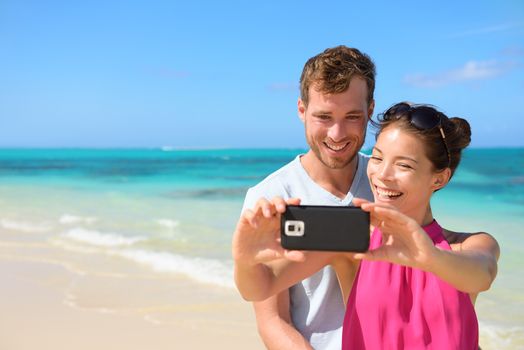 Smartphone - beach vacation couple taking selfie photograph using smartphone relaxing and having fun holding smart phone camera. Young beautiful multicultural Asian Caucasian couple on summer beach.