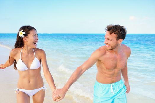 Beach couple having fun happy on beach vacation during summer holiday. Multiracial fit couple running together holding hands laughing in the sun. Young adults in shape carefree happiness.