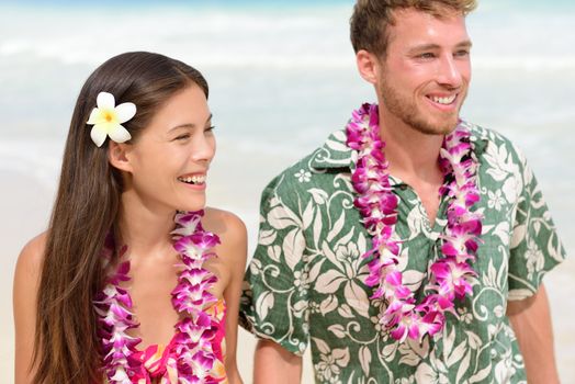 Happy Hawaii beach couple in Aloha Hawaiian shirt. Portrait of Asian woman and Caucasian man on beach walking with flower leis and typical attire for their wedding or honeymoon.