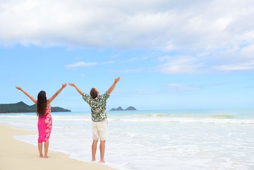 Happy freedom couple on Hawaiian beach vacations. Full length people standing with arms outstretched up to the sky showing happiness and success in Hawaii wearing traditional Aloha shirt and dress.