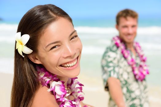 Happy Hawaii couple in Hawaiian lei, flower head and Aloha shirt. Portrait of a smiling Asian multiracial woman with Caucasian boyfriend on beach, multiethnic couple on summer travel vacations.