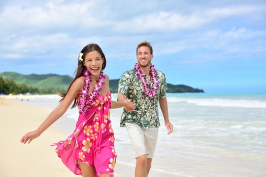 Happy couple having fun running on Hawaii beach vacations in Hawaiian clothing wearing Aloha shirt and pink sarong sun dress and flower leis for traditional wedding or honeymoon concept.