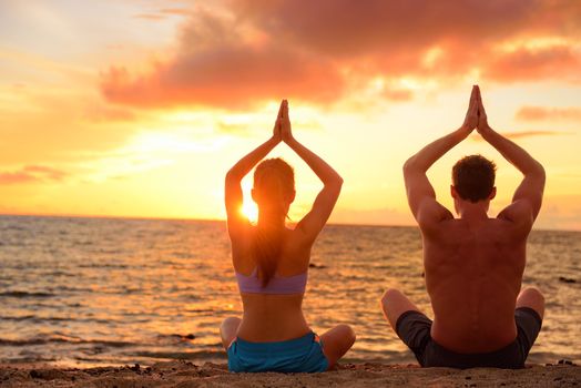 Yoga couple relaxing doing meditation on beach. Silhouettes of man and woman people practicing yoga pose sitting at a beach in the lotus position with their hands raised against a colorful sunset sky.
