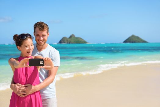 Beach vacation couple taking selfie photograph using smartphone, Lanikai beach, Oahu, Hawaii, USA with Mokulua Islands. Couple holding smart phone camera. Young beautiful multicultural Asian Caucasian