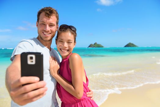 Smartphone - beach vacation couple taking selfie photograph using smartphone having fun holding smart phone camera on Lanikai beach, Oahu Hawaii, USA with Mokulua Islands. Young Asian Caucasian couple