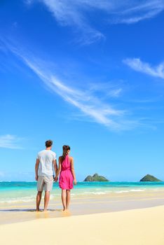 Beach vacation couple relaxing on summer holidays. Young people standing from behind holding hands looking at the ocean on Lanikai beach, Oahu, Hawaii, USA with Na Mokulua Islands.