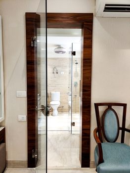 Glass doorway leading to a washroom with the commode visible and blue chair on the side Shot at a modern expensive beautifully maintained flat or apartment in Delhi, Gurgaon, Noida for premium individuals. Shows the real estate of India
