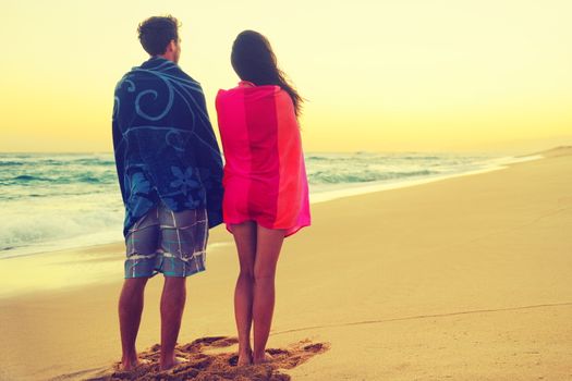 Romantic couple bathing with towels on beach sunset. Portrait of happy young couple looking at sunrise ocean sea view during holidays vacation travel. Asian woman, Caucasian man.