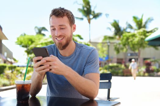 Man on cafe using smart phone app text messaging sms drinking iced coffee in summer. Handsome young casual man using smartphone smiling happy sitting outdoors. Urban male in his 20s.