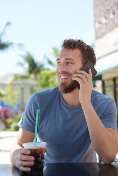 Man on cafe using smartphone talking on mobile cell smart phone drinking iced coffee in summer. Handsome young casual man using smartphone smiling happy sitting outdoors. Urban male in his 20s.