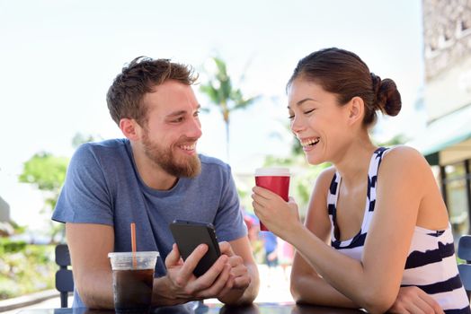 Couple on cafe looking at smart phone app pictures drinking coffee in summer. Young urban man using smartphone smiling happy to casual asian woman sitting outdoors. Friends in late 20s