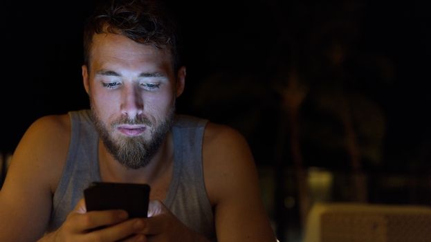 Man using smartphone at night browsing internet updating social media. Young male with beard using mobile cell smart phone outdoors in the dark in summer.