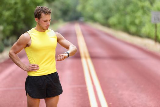 Runner looking at smartwatch heart rate monitor having break while running. Man jogging outside looking at sports smart watch during workout training for marathon. Fit male fitness model in his 20s.