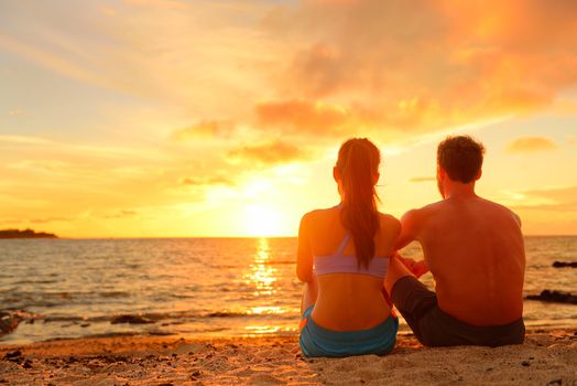 Happy Romantic Couple Enjoying Beautiful Sunset at the Beach Sitting in Sand looking at ocean sea and colorful yellow sky.