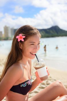 Beach woman drinking iced coffee cappuccino drink enjoying beach lifestyle smiling happy on Waikiki, Honolulu, Oahu, Hawaii, USA. Mixed race Asian Caucasian female in bikini.