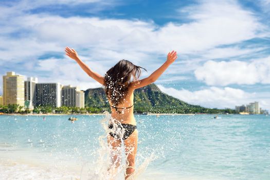 Beach fun - happy woman on Hawaii Waikiki vacation. Unrecognizable young adult from behind jumping of joy in water waves, arms up with diamond head mountain in the background, landmark of Honolulu.