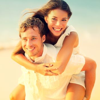 Romantic couple having fun piggyback on beach on honeymoon travel vacation summer holidays romance. Young happy lovers, Asian woman and Caucasian man doing playful joyful piggybacking ride outdoors.