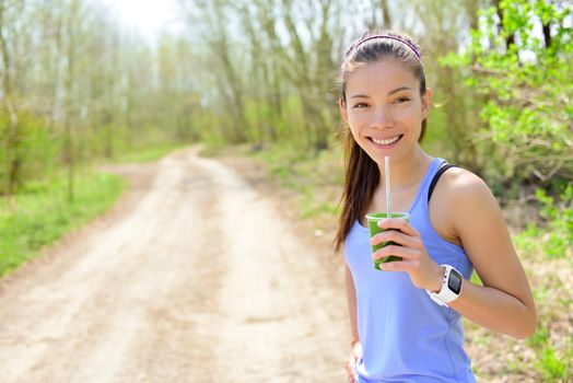 Healthy woman drinking green smoothie wearing smartwatch. Female runner resting drinking a spinach and vegetable smoothie using smart watch heart rate monitor during outdoor running workout in forest.