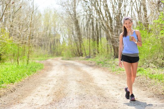 Woman runner drinking green smoothie wearing smartwatch. Female runner resting drinking a spinach and vegetable smoothie taking a break walking and resting during outdoor running workout in forest.