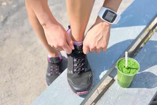 Running shoes, green vegetable smoothie and sports smartwatch. Female runner tying shoe laces in city park while drinking a healthy spinach and vegetable smoothie using smart watch heart rate monitor.
