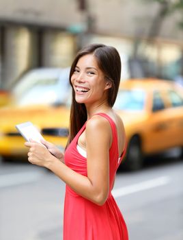 Woman in New York City using Tablet computer, standing in street with yellow taxi cab. Beautiful young woman in red dress smiling happy. Mixed race female model in Manhattan, New York City, USA.