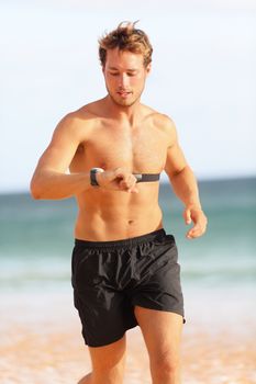 Male runner looking at heart rate monitor sports smart watch after running with earphones listening to music. Man athlete checking pulse during workout run exercise outdoors at ocean beach in summer.