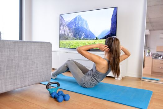 Home workout - woman exercising in front of a flat screen watching a fitness program or exercising during a TV show lying on a yoga mat in front of the sofa in the living room of a house or apartment.