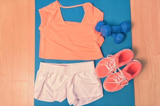 Workout clothes - fitness outfit and running shoes. Overhead of clothing ready for lifting weights at the gym or at home, laying on a yoga mat on the floor. Matching orange t-shirt and sneakers.