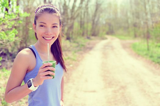 Healthy fitness girl drinking green spinach vegetable smoothie wearing smartwatch heart rate monitor during outdoor running workout in forest park during summer or spring. Happy fit Asian woman.