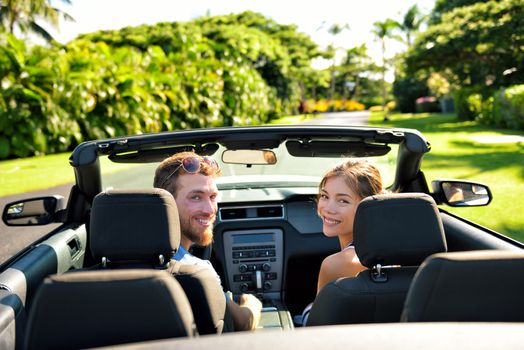 Happy couple in car on summer road trip travel. Multiracial young couple carefree on holidays driving a convertible cabriolet automobile on the roadway in the city looking back at camera.