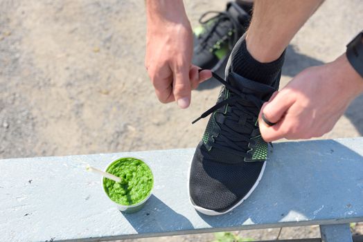Green smoothie and running - healthy lifestyle. Closeup of male runner's sport shoe tying laces on park bench for diet and weight loss concept for men. Getting ready for jogging and cardio workout.