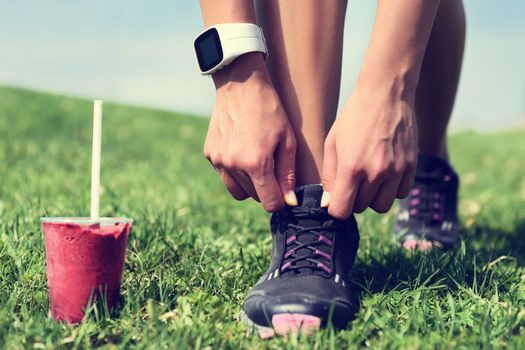 Fresh start on weight loss - runner tying laces with fruit smoothie wearing smartwatch for cardio. Detox, clean eating and diet. Woman getting ready for jogging workout. Closeup of running shoes.