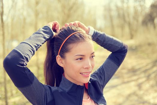 Woman tying hair in ponytail getting ready for run. Beautiful Asian young adult attaching her long brown hair on outdoor trail running path in forest to prepare for workout.