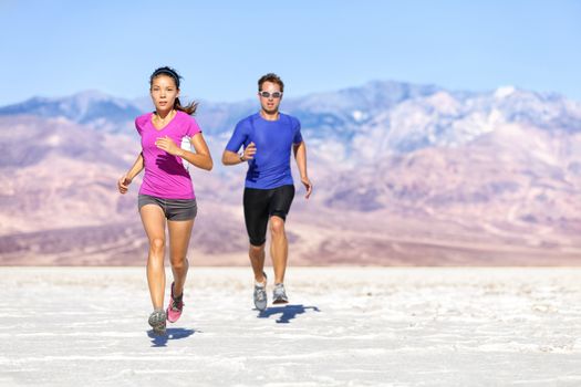 Runners trail running on dry desert landscape. Couple of fit athletes sprinting in compression activewear wearing sports clothing sweating in hot weather. Full length people in dramatic nature.