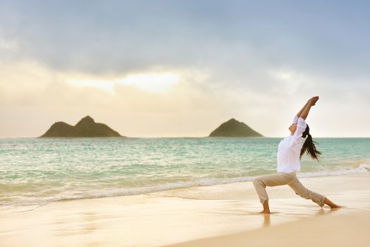 Yoga woman meditating in warrior pose relaxing outside on beach at sunrise. Female yoga girl working out training in serene ocean landscape. Lanikai beach, Oahu, Hawaii, USA.