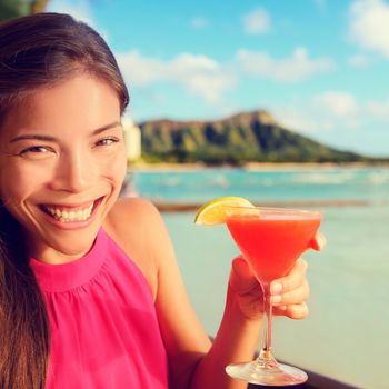 Woman drinking cocktail alcohol drinks at beach bar resort in Waikiki, Honolulu city, Oahu, Hawaii, USA. Asian girl tourist looking at camera toasting a glass of Hawaiian drink at sunset.