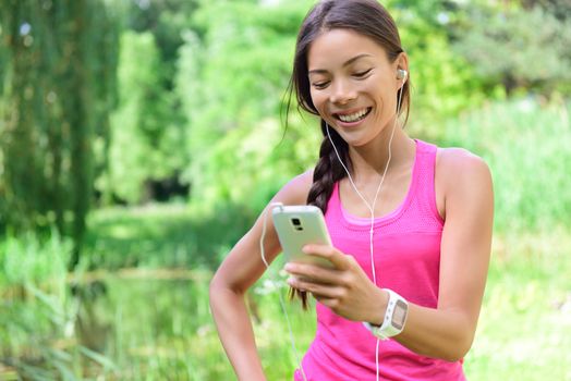 Woman runner sharing running data on social media after exercise. Girl listening to music on smart phone after jogging run in city park. Female jogger with earphones, smartphone and heart rate monitor.