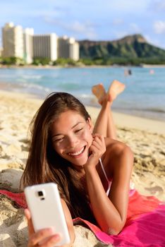 Smart phone woman using smartphone app on beach smiling laughing having fun. Girl reading or messaging or browsing on internet smiling happy outdoors. Mixed race female model on Waikiki Oahu, Hawaii.