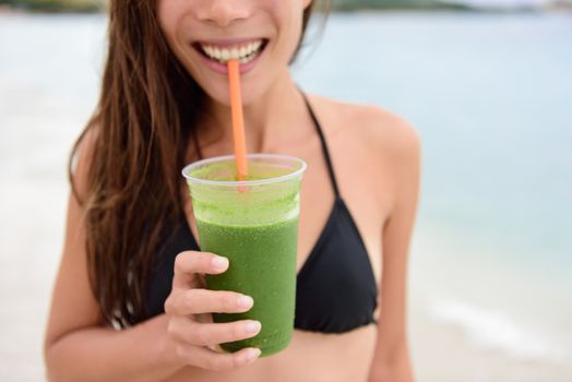 Green smoothie. Woman holding green vegetable detox juice outside in bikini in summer sun on beach. Healthy lifestyle with beautiful mixed race Asian Caucasian female model taking a cleanse diet.