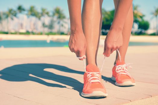 Low section and feet of fit young woman tying sports shoe lace. Mixed race Asian / Caucasian female jogger is on footpath going running. Her hands are tightening shoe laces on sunny day.