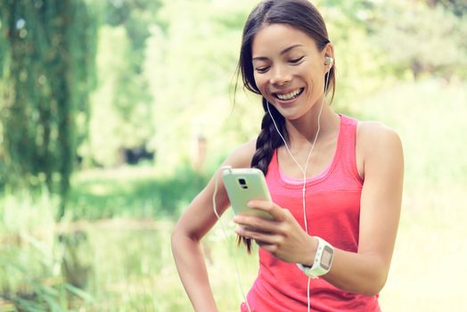 Happy young fit woman using cell phone while listening to music on smartphone. Smiling mixed race Asian / Caucasian female is in sports clothing. She is enjoying music in park.