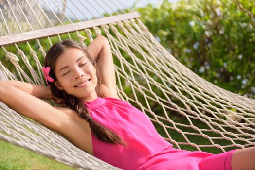 Happy relaxed young woman with hands behind head lying on hammock. Smiling mixed race Asian / Caucasian female is in casual summer dress. She is enjoying sunlight in park.