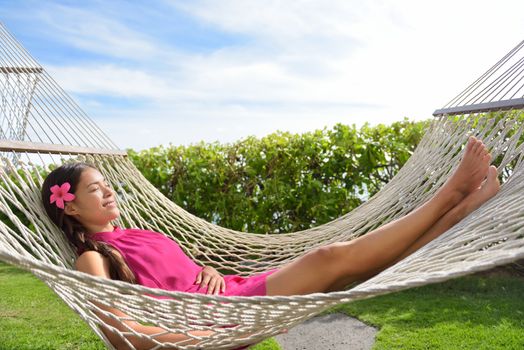 Full length of happy relaxed young woman lying on hammock. Smiling mixed race Asian / Caucasian female is in dress. She is enjoying sunlight in park.