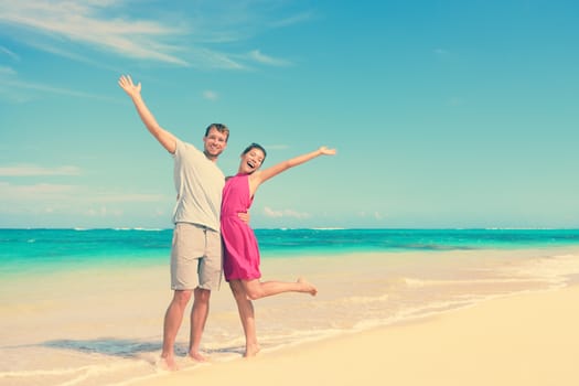Full length portrait of happy couple with arms raised standing on shore. Joyful partners are enjoying at beach. Carefree tourists are celebrating summer vacation in idyllic nature.